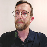 cable-4-leitung-technik-benjamin-viola Mann mit braunem Bart, Brille und dunkelblauem Poloshirt vor hellem Hintergrund, frontal und leicht lächelnd, ideal für professionelle Produktpräsentationen oder Teamseiten im Onlineshop, freundlicher und seriöser Ausdruck.