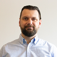 cable-4-service-team-sebastian-wozniak Mann mit dunklem, kurzem Haar und Vollbart trägt ein hellblaues Hemd, steht vor hellem, neutralem Hintergrund und blickt freundlich in die Kamera. Ideal für professionelle Profilfotos oder Business-Darstellung